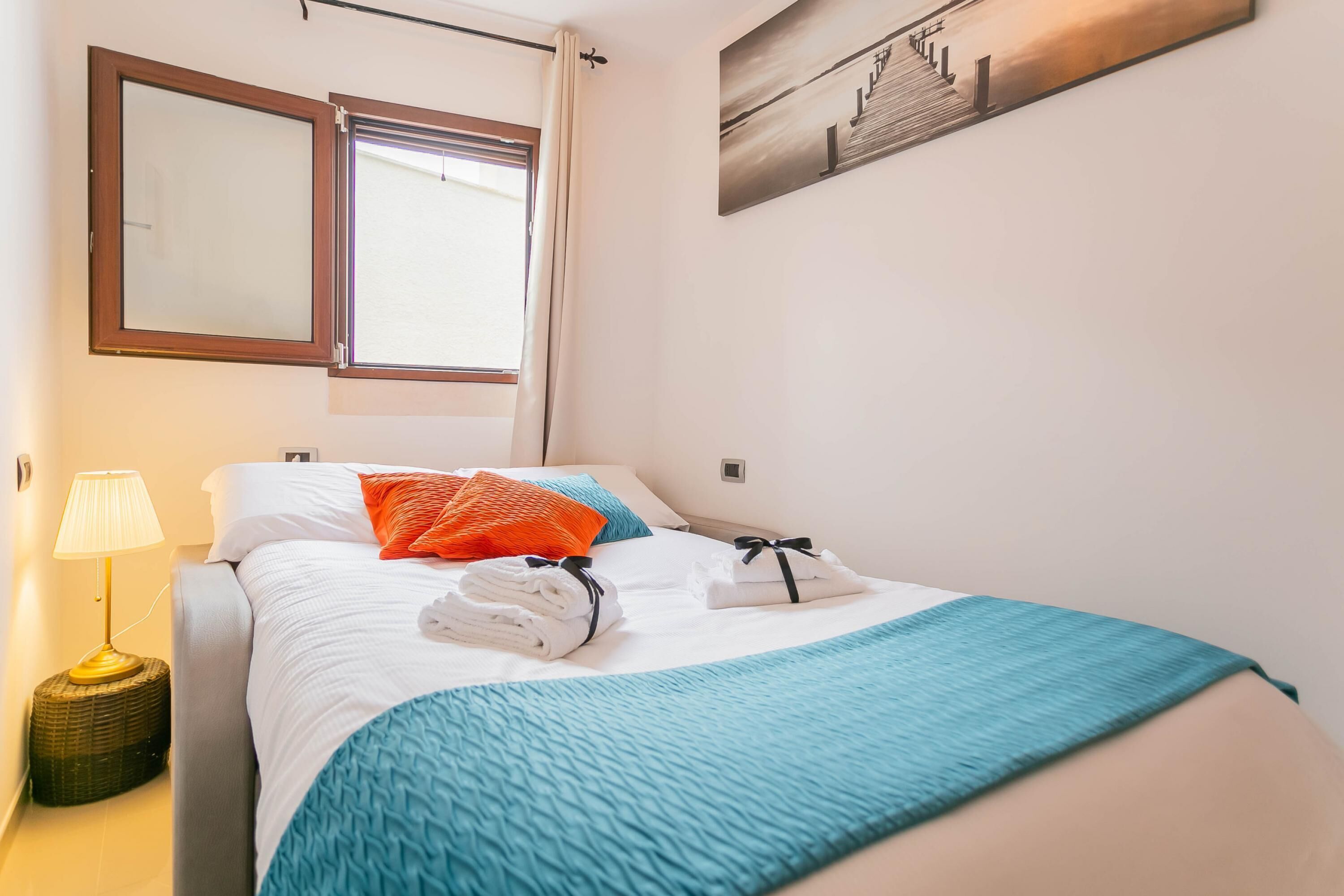 Ein ordentlich bezogenes Bett mit weißer und türkisfarbener Bettwäsche, ein Fenster mit Vorhängen, eine Lampe und ein Bild an der Wand.