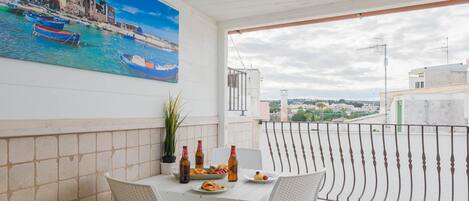A balcony with a table set for two, a potted plant, and a painting of a coastal scene.
