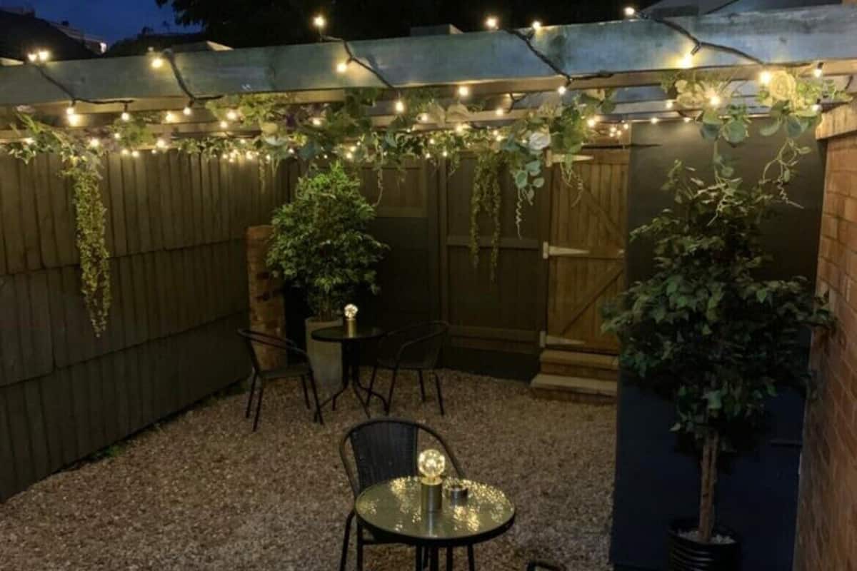 An outdoor patio area with a gravel floor, surrounded by wooden fencing, featuring string lights and potted plants.
