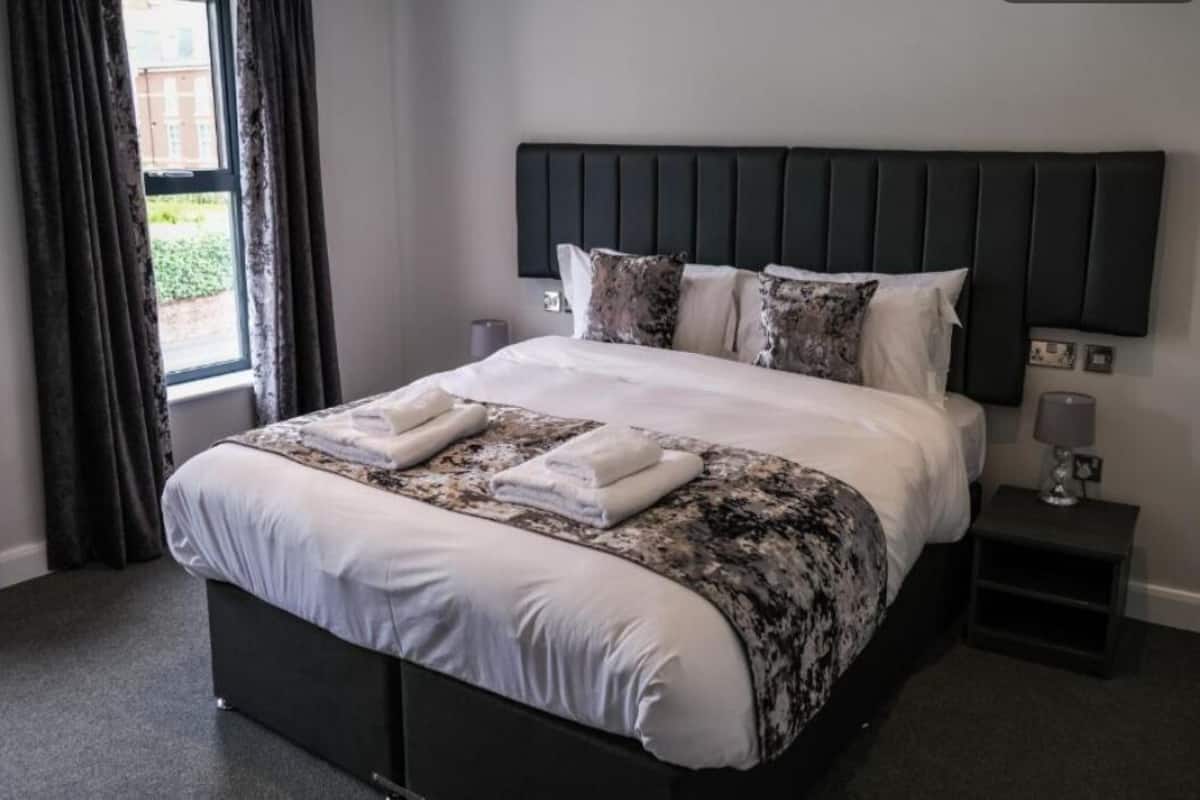 A neatly made bed with a dark headboard, a bedside table with a lamp, and a window with curtains.