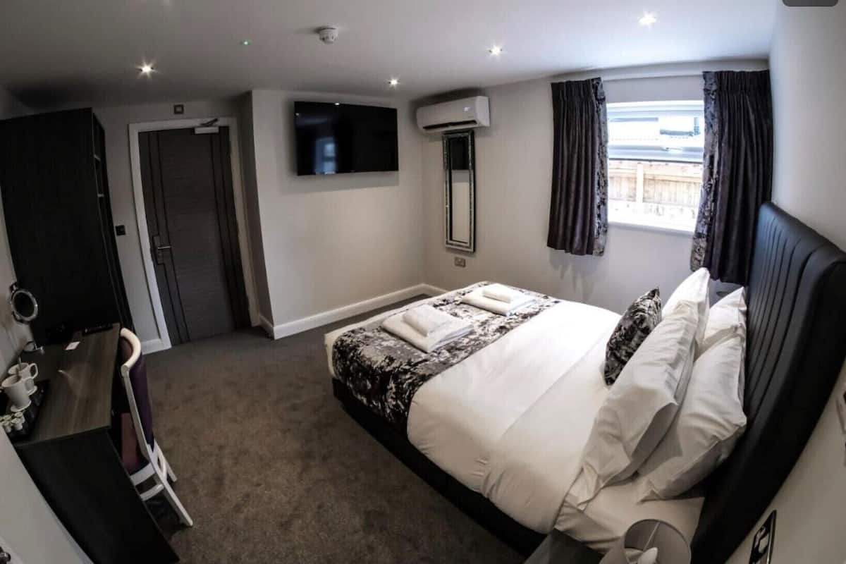 A hotel room with a bed, a TV mounted on the wall, an air conditioning unit, a window with curtains, and a door.