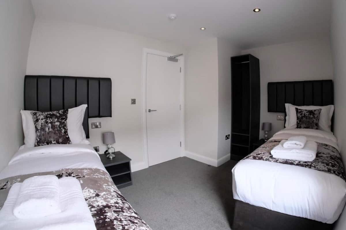 A hotel room with two beds, each with a dark headboard and white bedding. There are bedside tables with lamps, a door, and a wardrobe.