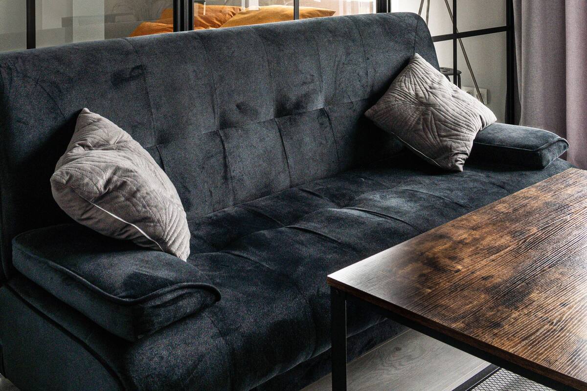 Een moderne woonkamer met een donkergrijze bank, twee kussens, een houten salontafel en een groot raam met gordijnen.