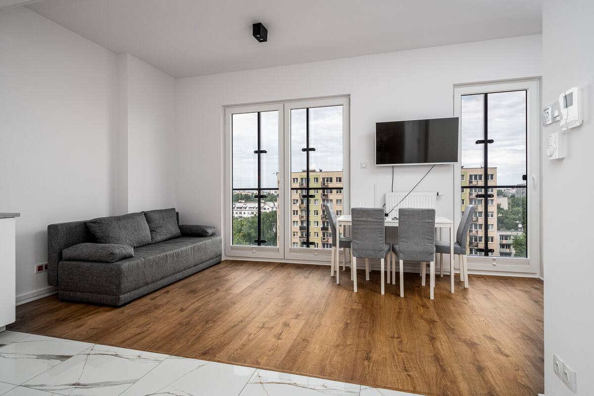 Een moderne woonkamer met een grijze bank, een eettafel met stoelen, een aan de muur bevestigde televisie en grote ramen met uitzicht op de stad.