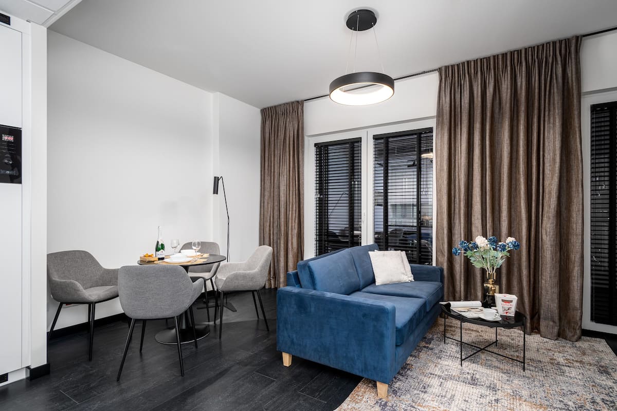 Een moderne woonkamer met een blauwe bank, grijze fauteuils, een eettafel met stoelen en een groot raam met gordijnen.