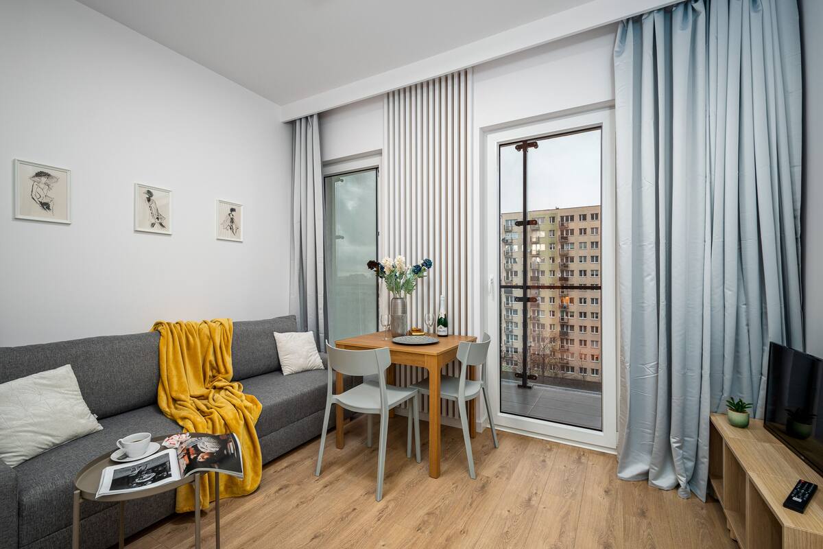 Een moderne woonkamer met een grijze bank, een houten eettafel met twee stoelen, een tijdschrift op een bijzettafeltje en een raam met gordijnen.