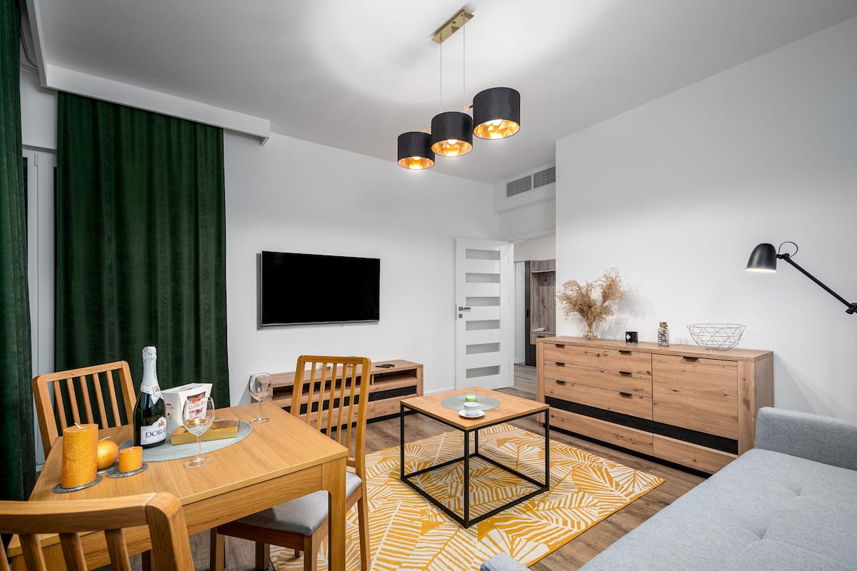 Een moderne woonkamer met een houten eettafel, stoelen, een bank, een televisie, een kast en een kamerbreed tapijt met een patroon.