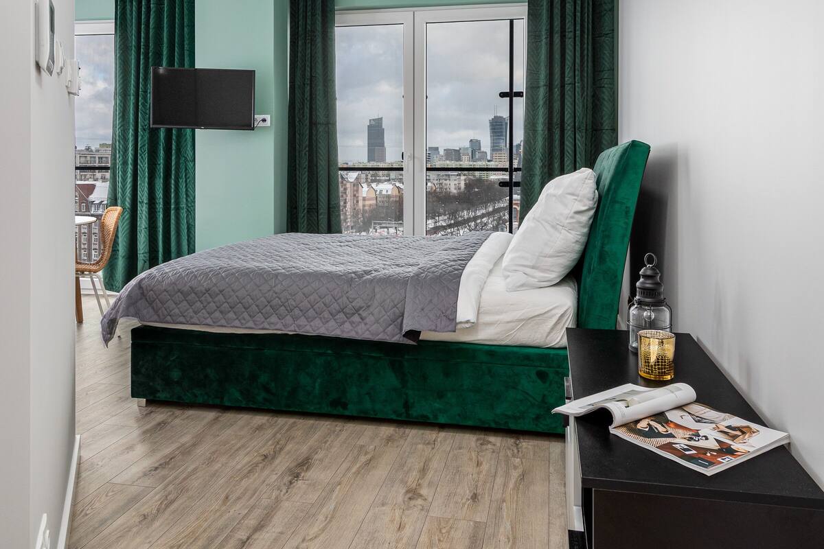 Een moderne slaapkamer met een bed, groene gordijnen, een raam met uitzicht op de stad, een flatscreen-tv en een klein tafeltje met een tijdschrift.