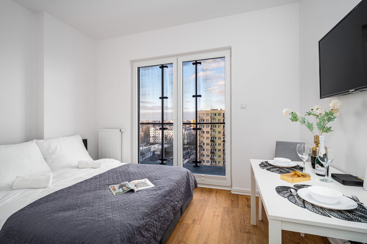 Een moderne hotelkamer met een groot bed, een eettafel gedekt voor twee personen en uitzicht op de stad door middel van kamerhoge ramen.