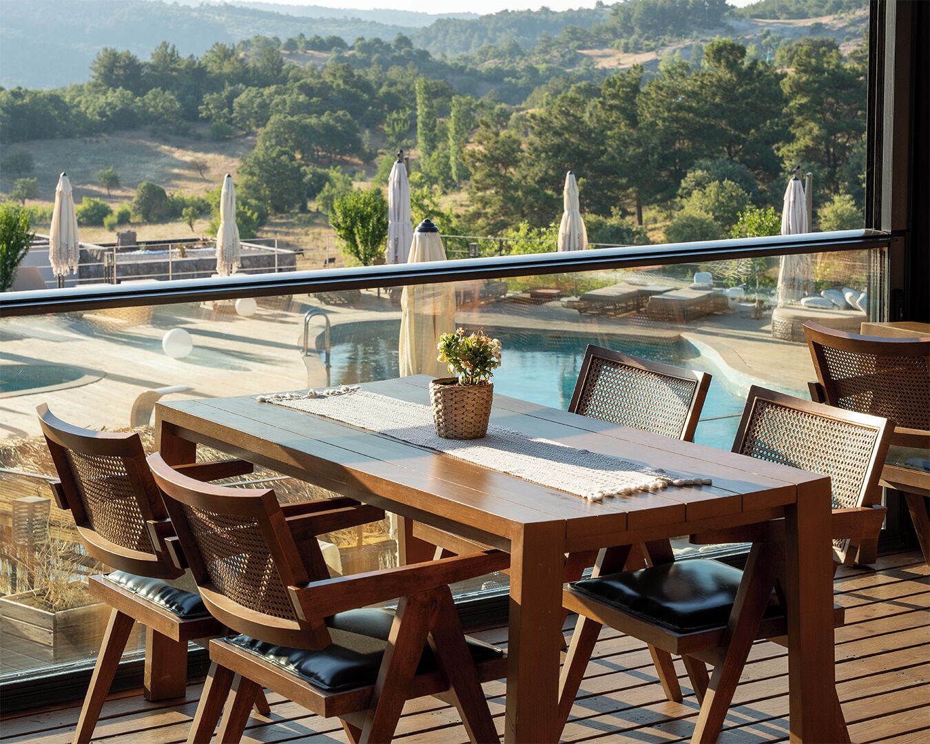 Una terraza de madera con una mesa y sillas, con vista a una piscina y abundante vegetación.