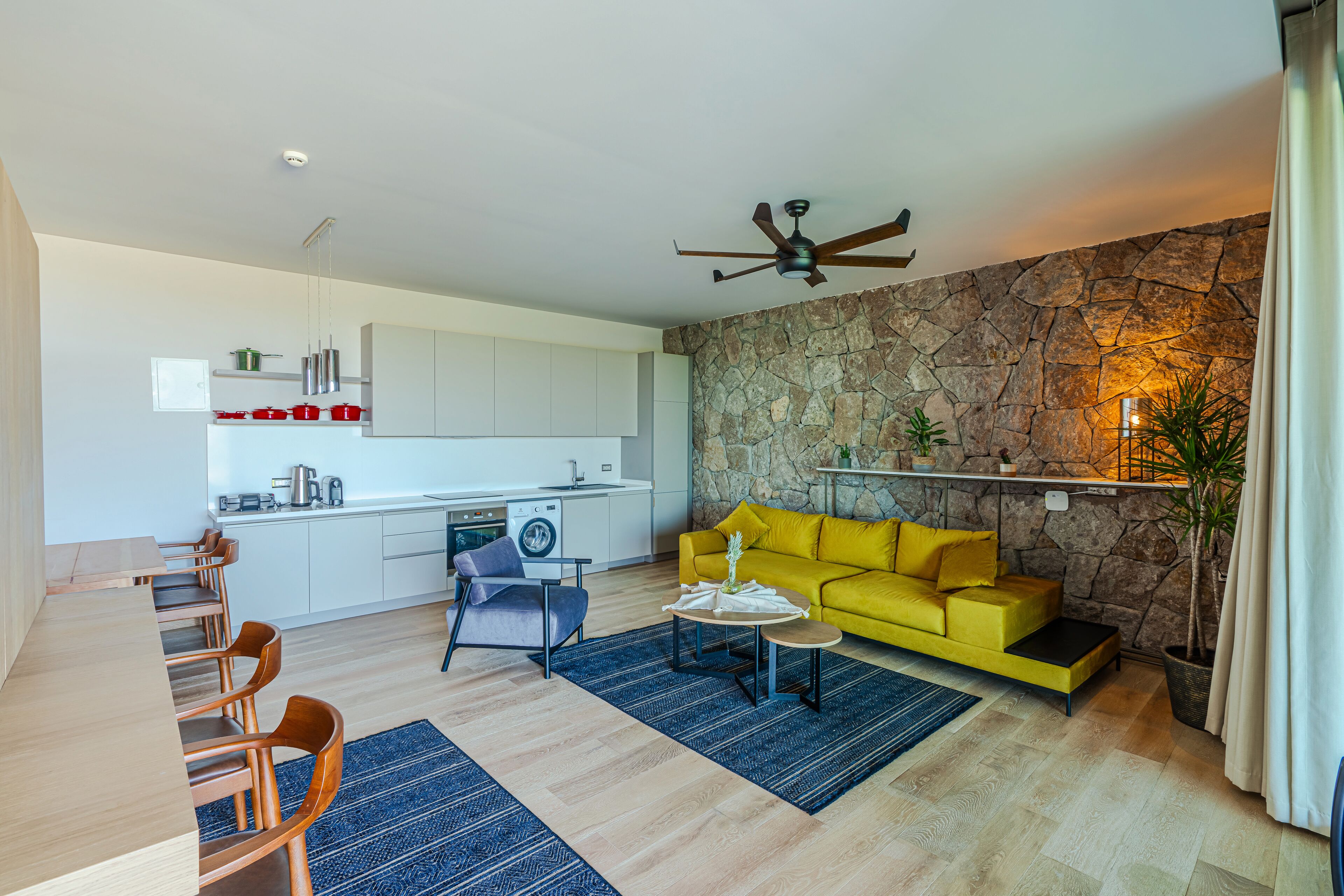 A modern living room with a yellow sofa, a dining area with wooden chairs, a kitchen with white cabinets, and a stone accent wall.