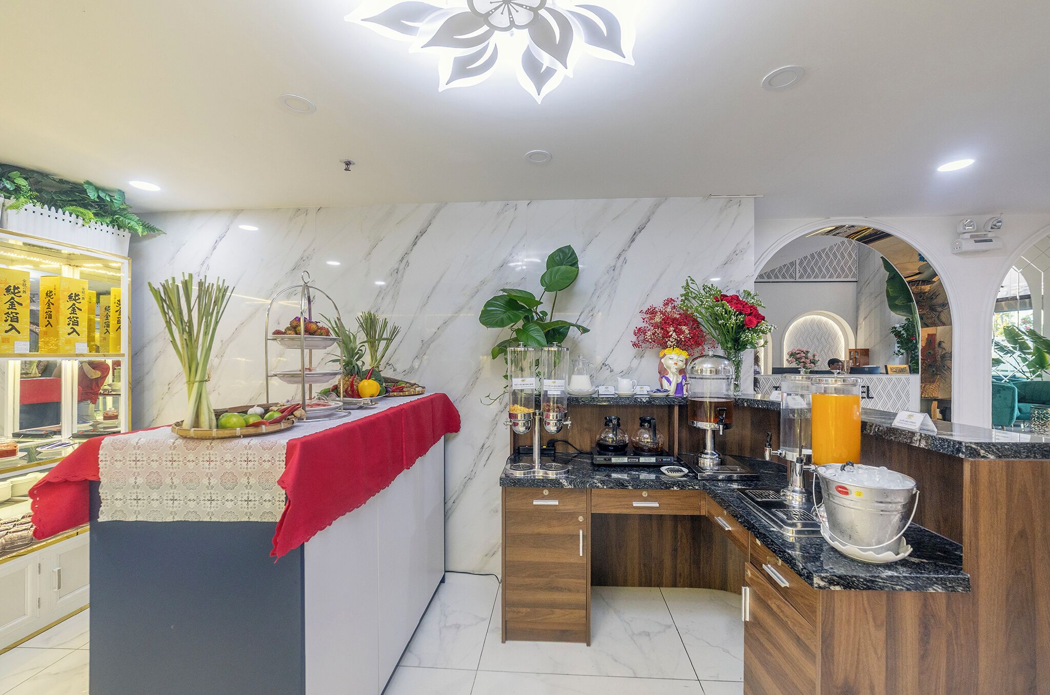 A modern kitchen with marble countertops, wooden cabinets, and a red tablecloth. There are plants, a fruit bowl, and a coffee machine.
