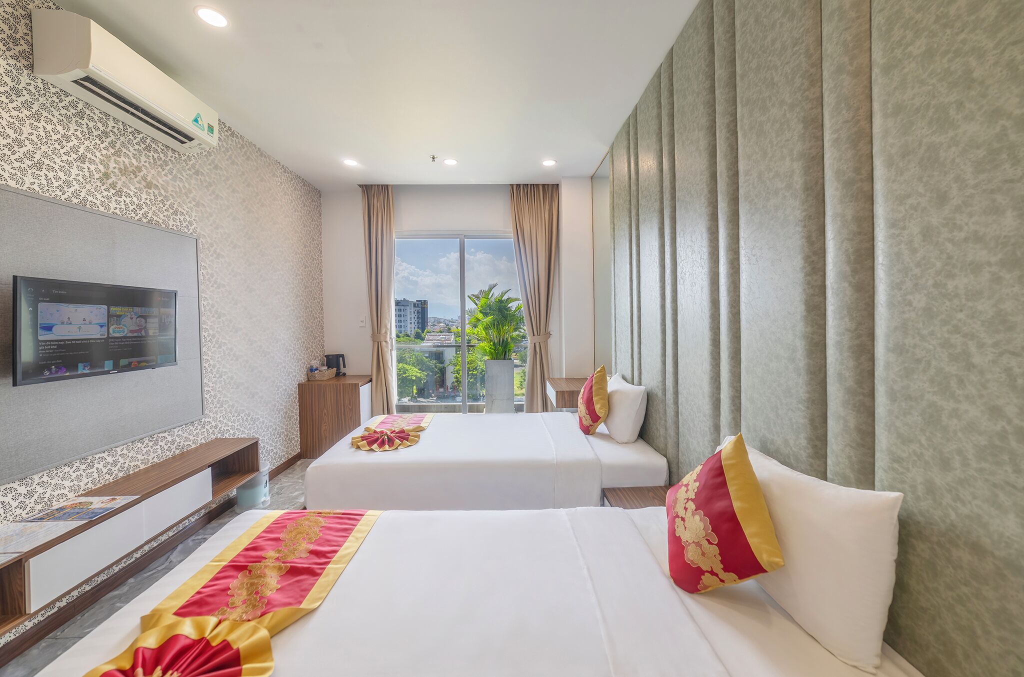 Une chambre d’hôtel avec deux lits, une télévision, une grande fenêtre avec des rideaux et une vue sur les bâtiments et la verdure environnante.