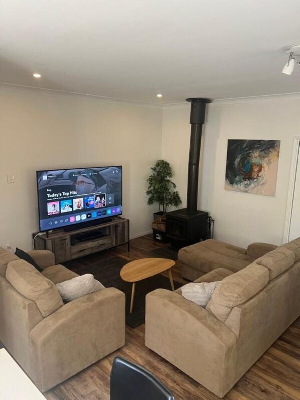 A living room with a flat-screen TV, a wooden coffee table, beige sofas, a potted plant, and a black fireplace.