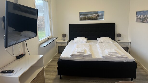 A hotel room with a large bed, a flat-screen TV mounted on the wall, a window with a view of the outdoors, and a painting on the wall. - Gæstehus (Grasten)