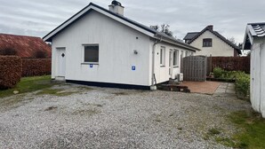A white building with a single window and a door, a gravel driveway, and a wooden fence. - Gæstehus (Grasten)