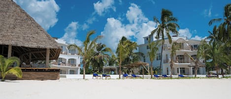 A beachfront hotel with thatched-roof structures, palm trees, and blue lounge chairs.