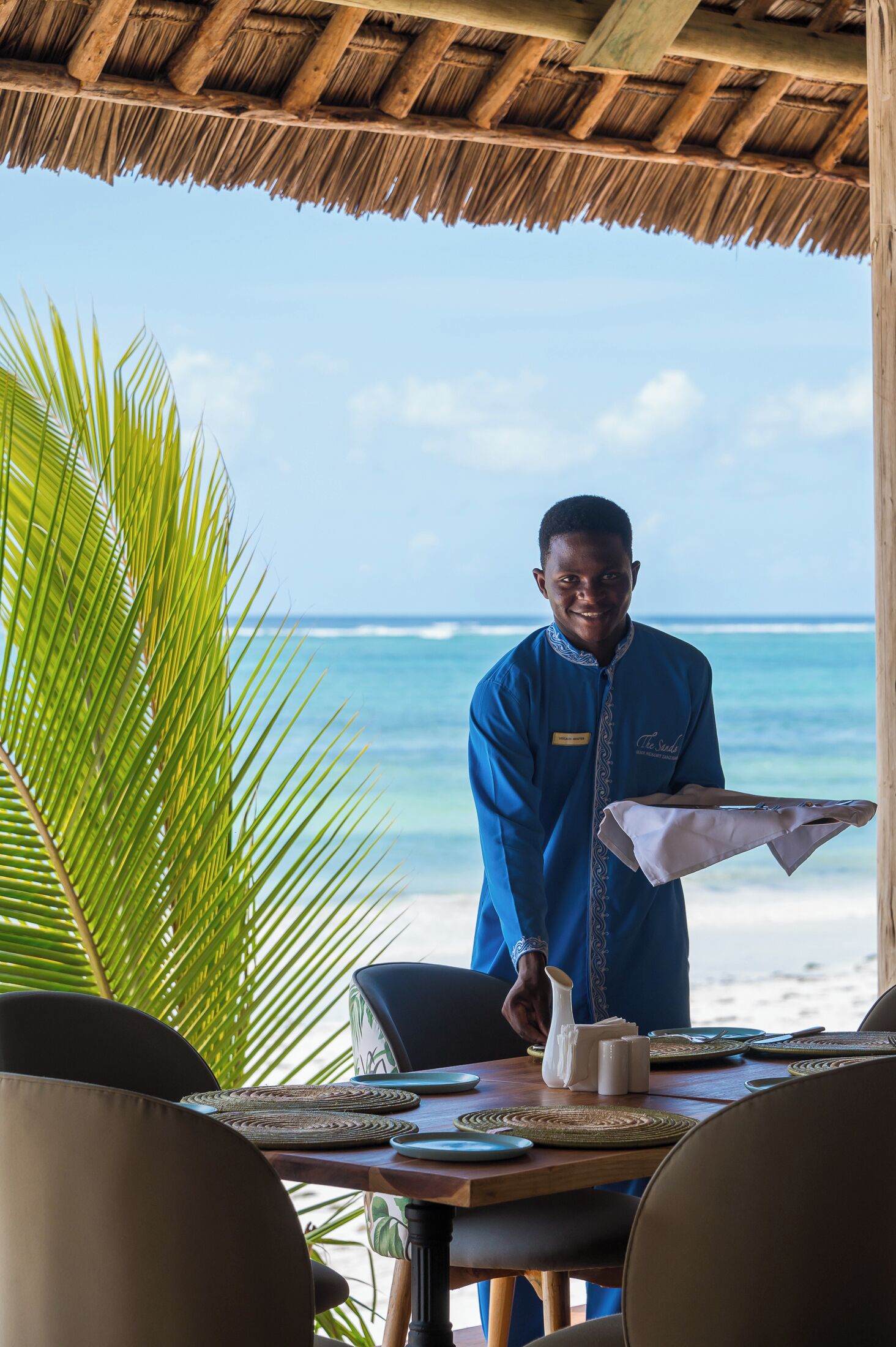 Eine Person in blauer Uniform steht an einem Tisch mit Blick auf den Strand, Palmenblättern und einem Reetdach.