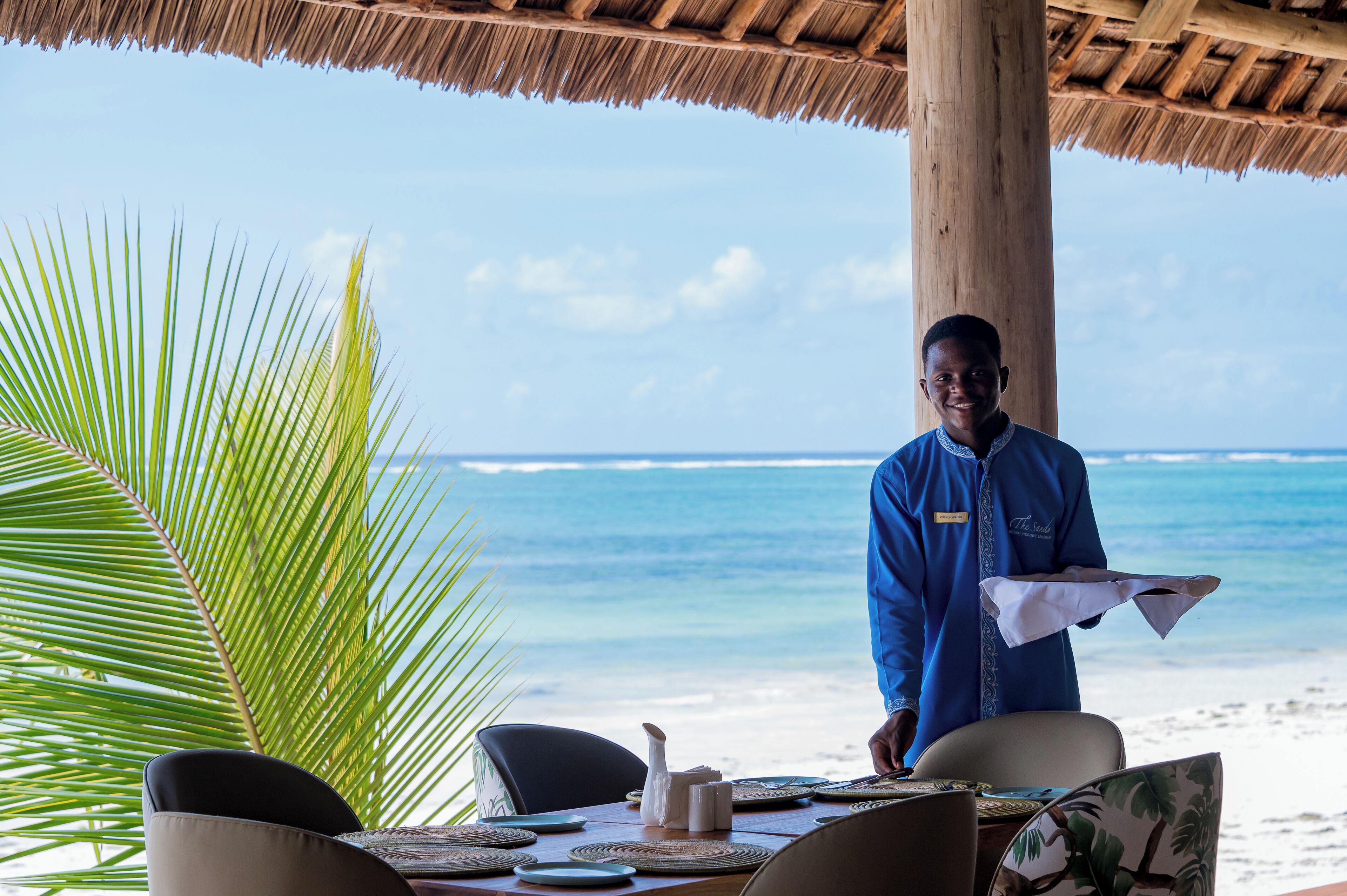 A beachside dining area with a table set for two, a palm tree, and a view of the ocean.
