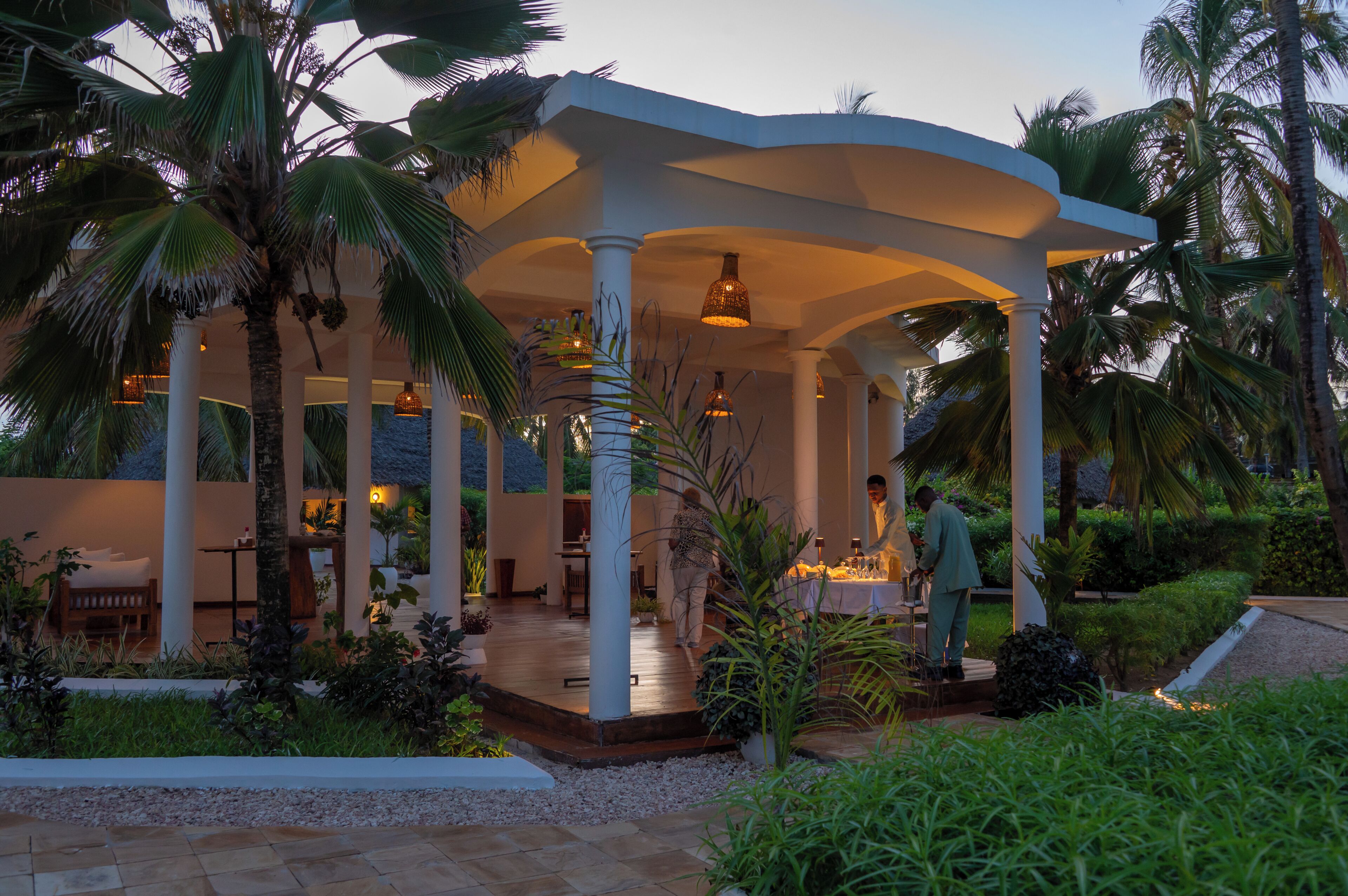 A covered outdoor area with a dining setup, palm trees, and a white structure.