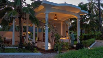 A covered outdoor area with a dining setup, palm trees, and a white structure.
