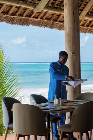 A person in a blue uniform serving food at a wooden table with a view of the ocean. - Ocean Boutique Hotel (Dongwe)