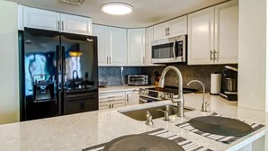 A modern kitchen with white cabinets, a stainless steel refrigerator, a microwave, and a double sink with a striped mat. - Oceanfront Modern Condo w 8 Water Features (Myrtle Beach)
