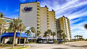 A hotel with multiple floors, a prominent sign, and a parking area with cars. - Oceanfront Modern Condo w 8 Water Features (Myrtle Beach)
