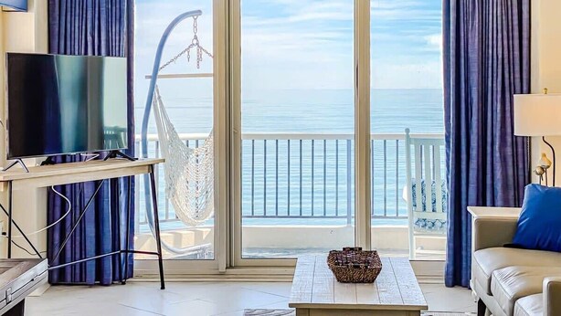 A room with a TV on a stand, a white table with a bowl, a sofa, and a chair. There is a large window with curtains, a hammock, and a view of the sea. - Oceanfront Modern Condo w 8 Water Features (Myrtle Beach)