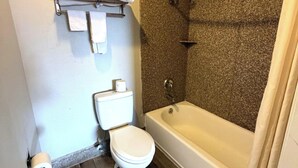 A bathroom with a toilet, a roll of toilet paper, a towel rack, and a bathtub. - Regency Inn Three Rivers I-37 (Three Rivers)