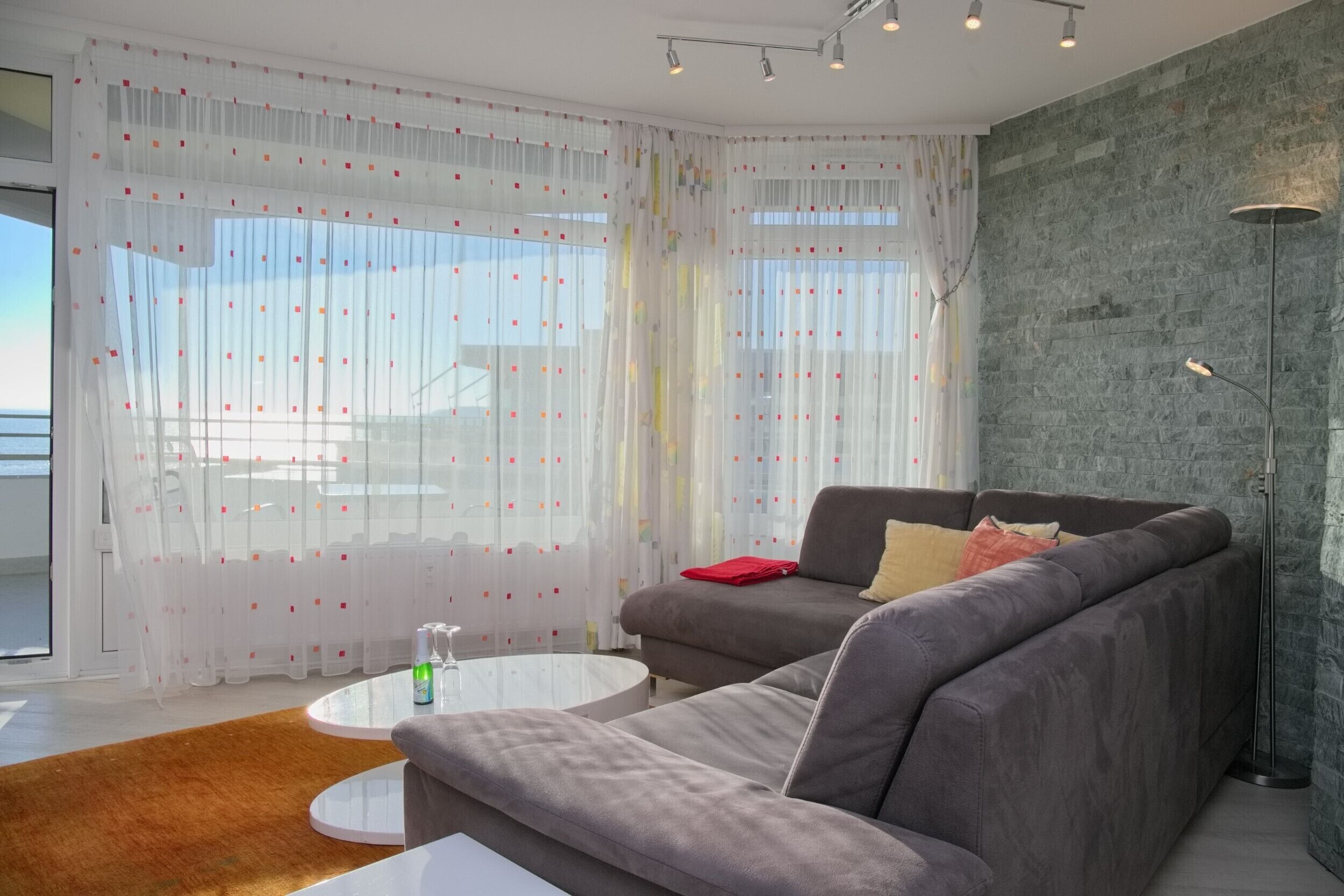 A modern living room with a grey sofa, a white coffee table, and a red throw pillow. There is a large window with sheer curtains and a view of the outside.