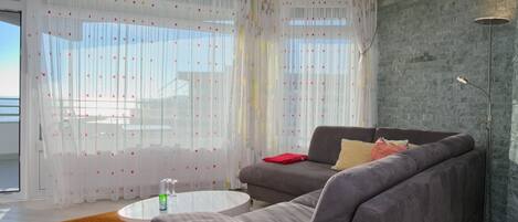A modern living room with a grey sofa, a white coffee table, and a red throw pillow. There is a large window with sheer curtains and a view of the outside.