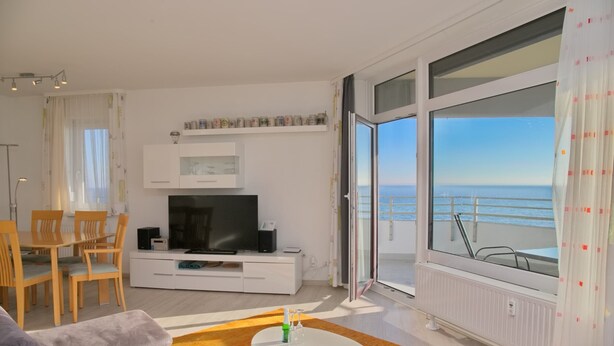 A modern living room with a TV, dining area, and a large window with a view of the sea.