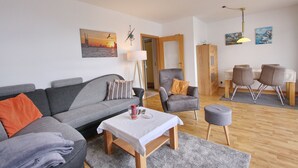 A modern living room with a grey sofa, a small table, a wooden chair, and a dining area with a table and chairs. - Am Brandungsbad App. 3.2 (Grömitz)