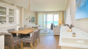 A modern living room with a wooden dining table, grey upholstered chairs, a white sofa, and a large window offering a view of the sea.