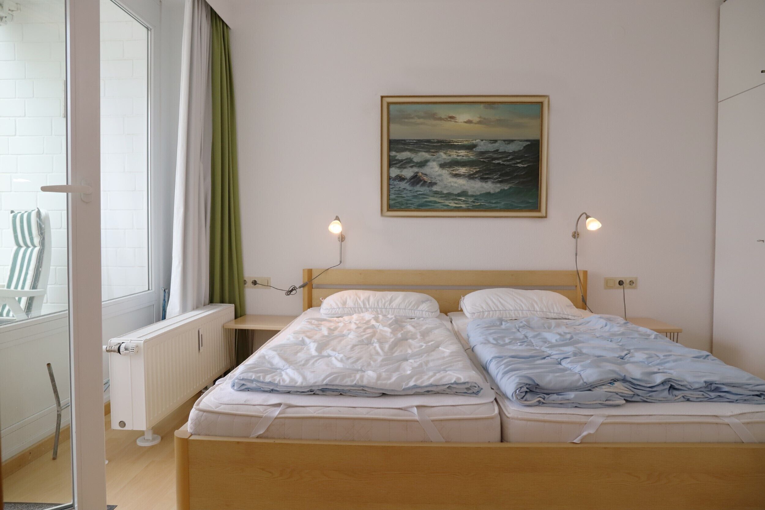 A double bed with white and blue bedding, two bedside lamps, a painting of the ocean, and a window with curtains.