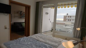 A bedroom with a bed, a bedside lamp, a window with a view of buildings, and a door leading to a balcony. - Tannenburg App. 504 Inkl. Wlan (Grömitz)