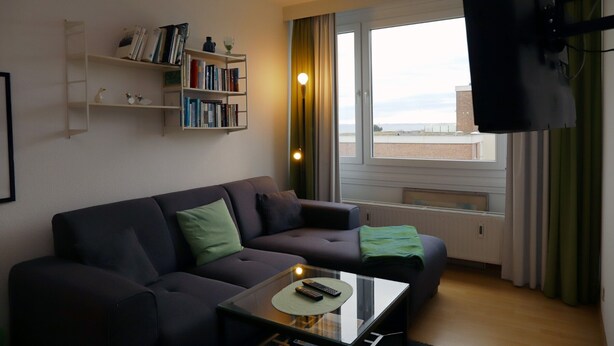 A modern living room with a sofa, a glass coffee table, a bookshelf, and a large window with curtains. - Tannenburg App. 504 Inkl. Wlan (Grömitz)
