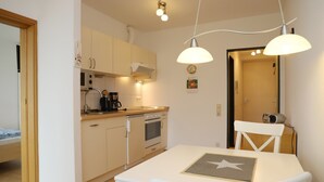 A compact kitchen with white cabinets, a stove, and a coffee maker. There is a dining table with a star placemat and white chairs. - Tannenburg App. 504 Inkl. Wlan (Grömitz)