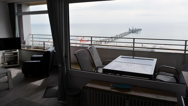Un balcón con vista al mar y un muelle. Hay una mesa, sillas, un televisor y un radiador.