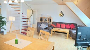 A modern living room with a wooden dining table, a sofa, a TV, and a staircase. - House Deichgraf Whg. 12 (Grömitz)