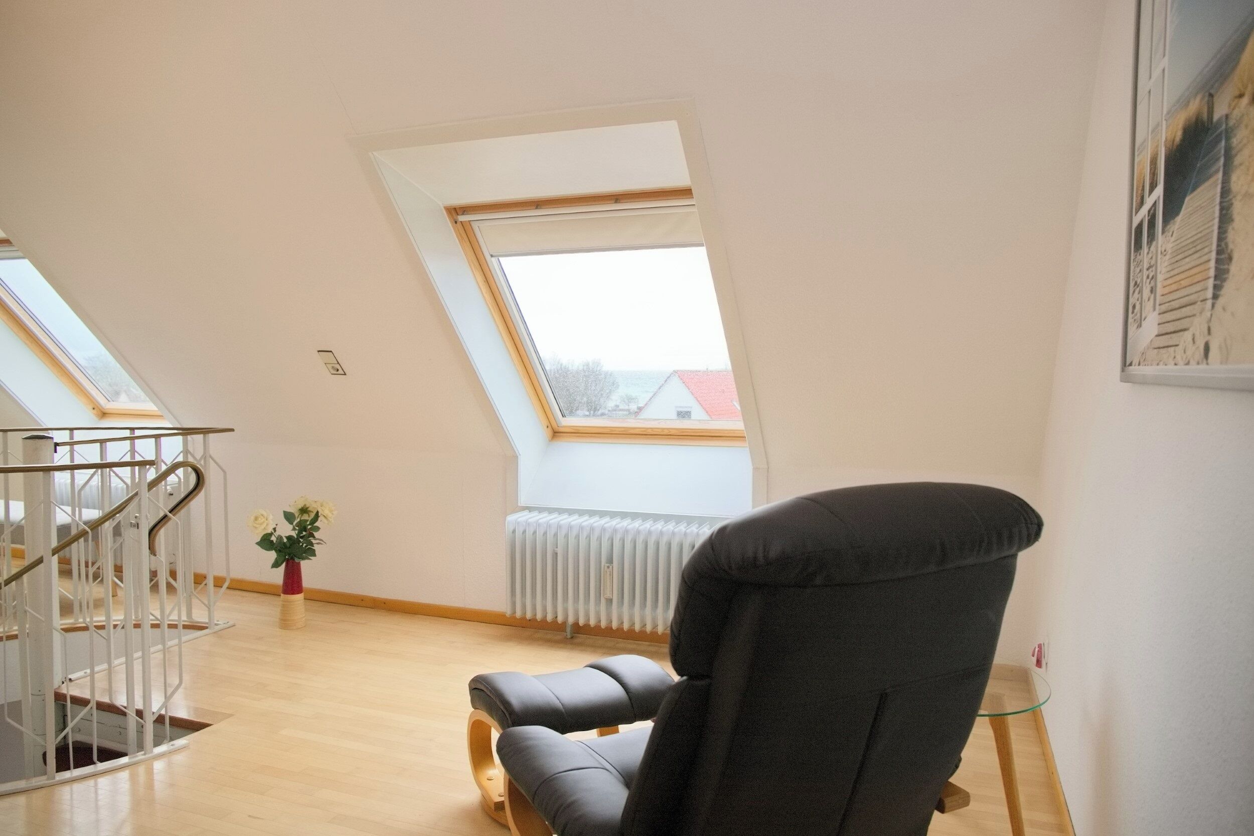 A room with a sloped ceiling, a radiator, a black chair, a small table, a staircase, and a window with a view of a building.