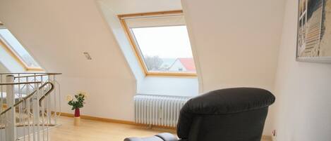 A room with a sloped ceiling, a radiator, a black chair, a small table, a staircase, and a window with a view of a building.