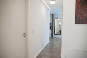 A hallway with a door on the left, a light fixture on the ceiling, and a mirror on the right wall.