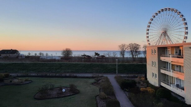 A modern building with a large Ferris wheel in the background, a grassy area with pathways, and a view of the sea. - Haus Baltic App. 188 Inkl. Wlan (Grömitz)
