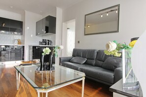 Un salon moderne avec une table basse en verre, un canapé gris et un coin cuisine à l’arrière-plan.