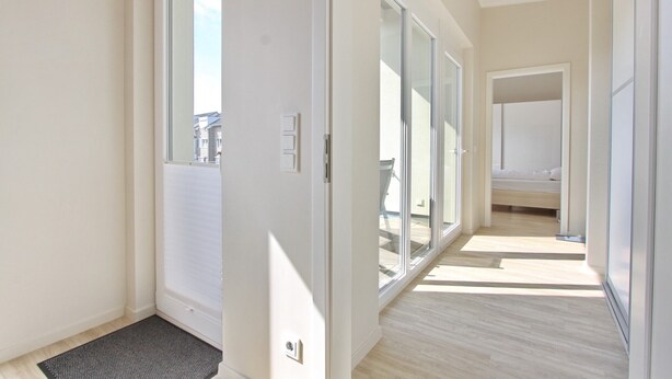 A bright hallway with a sliding glass door leading to a balcony, a bed in the background, and a wooden floor.
