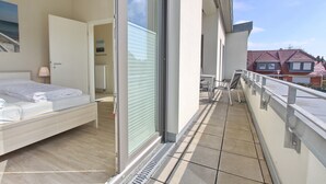 A bedroom with a bed, a sliding glass door leading to a balcony, and a view of red-roofed houses in the background.