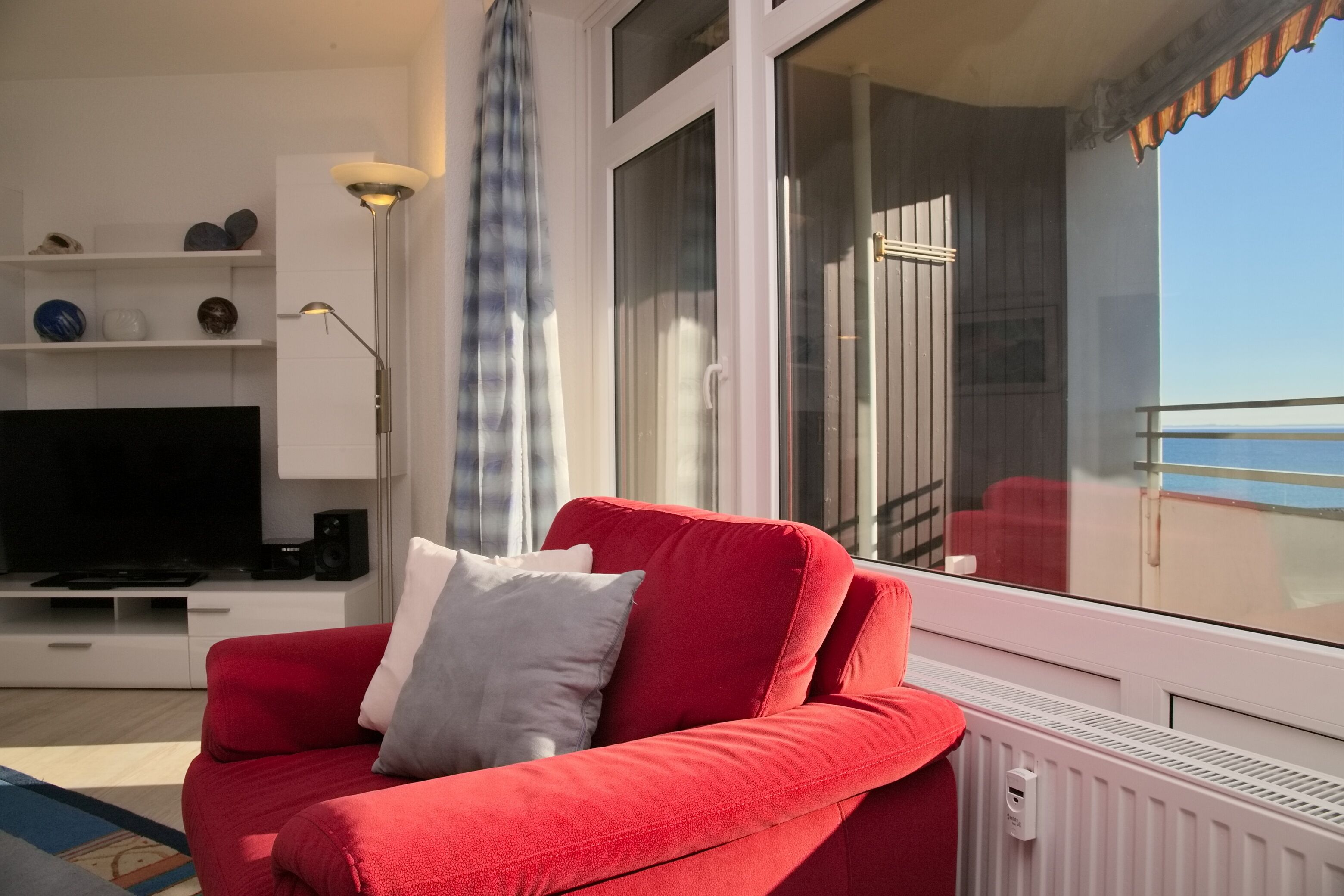 Un divano rosso con un cuscino bianco, una TV e un mobiletto bianco in un soggiorno con vista sul balcone.