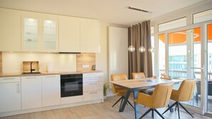 A modern kitchen with white cabinets, a wooden countertop, and a black dishwasher. There is a dining area with a wooden table and yellow chairs. - Brookgang 47d Doppelhaushälfte Inkl. Wlan (Grömitz)